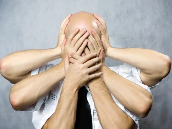 A bald, white man facing the camera with three pairs of white hands covering his face.